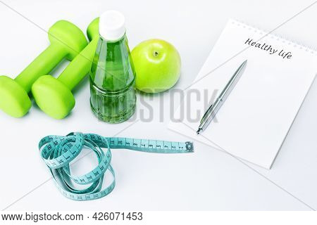Healthy Life Written In A White Notebook Near The Apple, Bottle Of Juice And Sport Equipment On A Wh