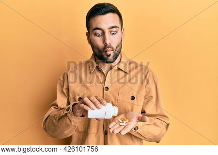 Young hispanic man holding pills making fish face with mouth and squinting eyes, crazy and comical. 