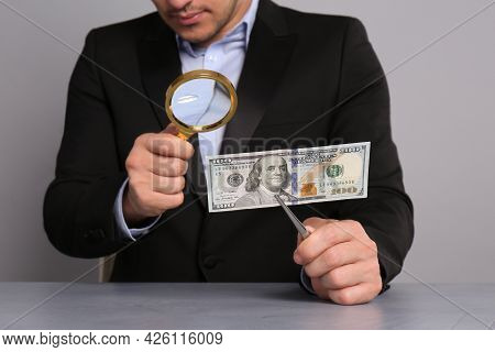 Expert Authenticating 100 Dollar Banknote With Magnifying Glass At Table On Light Grey Background, C