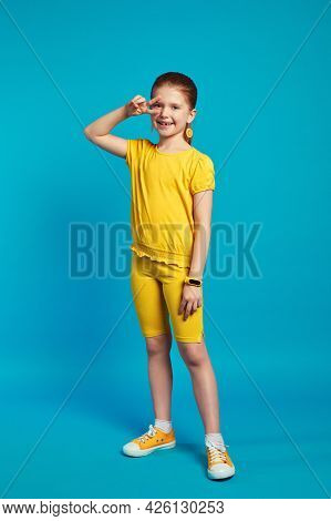 Vertical Full Body Shot Of Cute Little Girl In Yellow Outfit Smiling And Keeping Two Fingers Near Ey