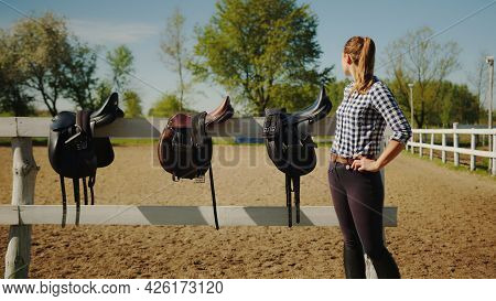 Leather Saddles Hanging On The Wooden Fence In The Sandy Arena. Girl In The Foreground Looking Back 