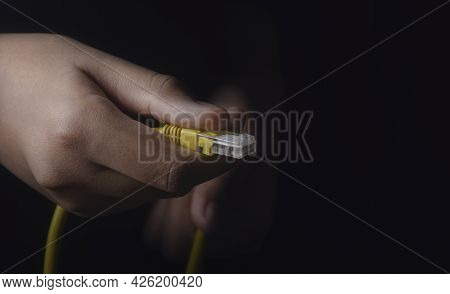 Front Hand Holding A Yellow Lan Ethernet Cable, And Back Hand Out Of Focus On The Black Ground. The 