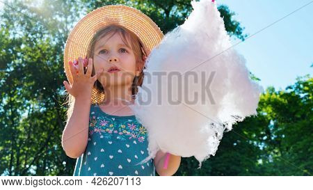 The Girl Looks In Amazement And Surprise At The Amusement Park. A Child Eats Cotton Candy On A Sunny