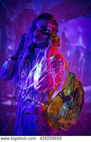 A cyberpunk blonde girl in a neon shiny raincoat, with a backpack and headphones stands on the street of the night cyber city of the future. Game, virtual reality. 