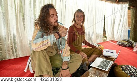 Young Couple Of Hippies Sitting In A Tent And Lights Up A Joint, Man Wears Beige Pants And Shirt In 