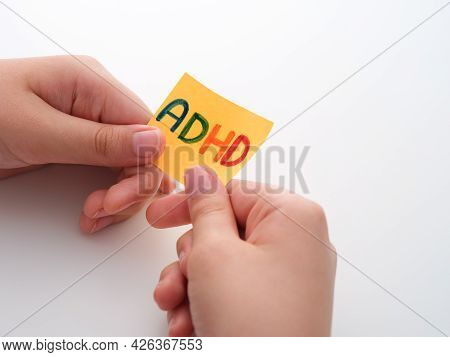 Adhd. A Child Holding A Yellow Paper Note In His Hands With The Abbreviation Adhd On It. Close Up. A