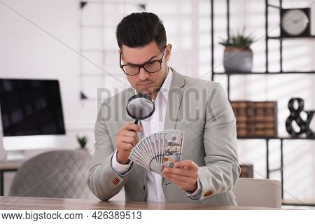 Expert Authenticating 100 Dollar Banknotes With Magnifying Glass At Table In Office. Fake Money Conc