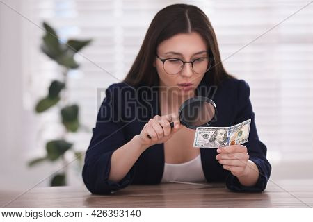 Expert Authenticating 100 Dollar Banknote With Magnifying Glass At Table In Office, Focus On Hand. F