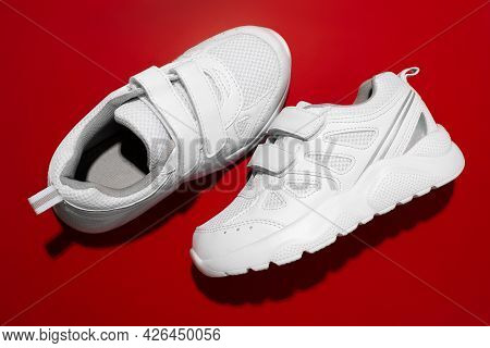 Top View Two White New Childrens Sneakers With Velcro For Easy Footwear On A Red Background, One Sne