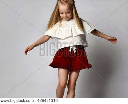 Little Blonde Girl Dances And Whirls On A White Background. Child In A White T-shirt And Red Shorts 