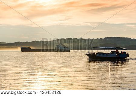 Passenger Boat And A Cargo Barge Float Down A Wide River In The Evening Light