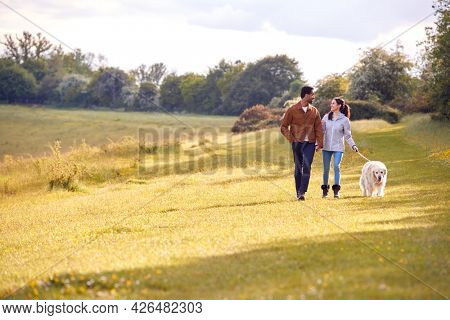 Couple With Pet Golden Retriever Dog Walking Along Path Across Field In Countryside