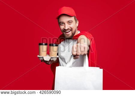 Positive Male Courier In Red Superhero Cloak And Cap Carrying Paper Bag And Disposable Cups Of Coffe