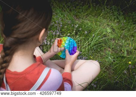A Little Cute Girl Is Sitting On The Grass With A Pop It - Antistress Toy.