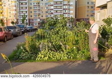 A Gardener In Overalls With A Yellow Hose Watering Flowers Against The Background Of Apartment Build