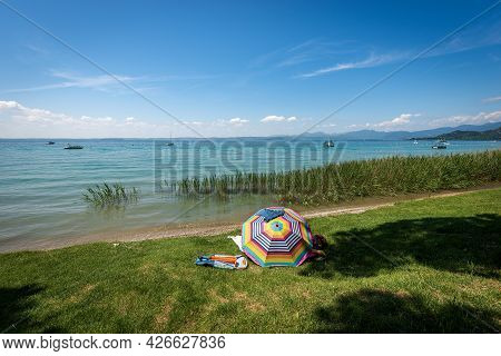 Beautiful Beach With Green Lawn And A Multi Coloured Umbrella On Lake Garda (lago Di Garda) Between 
