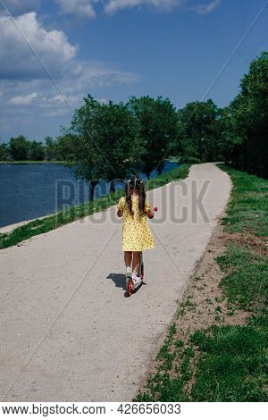 Lifestyle Portrait Of A Depersonalized Girl Riding A Scooter Along The River In The Park On A Family