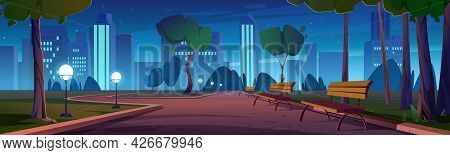 City Park With Wooden Benches, Green Trees And Grass, Lanterns And Town Buildings On Skyline At Nigh