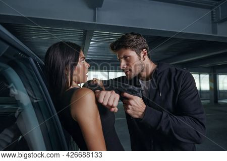 Aggressive robber threatening with pistol to frightened girl. European man and young brunette woman stand near auto. Male bandit wear hoodie. Concept of robbery and kidnapping. Inside parking lot