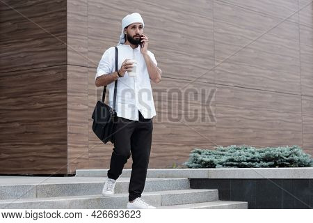 Pensive young bearded Muslim businessman in ghutrah moving down stairs and talking by phone