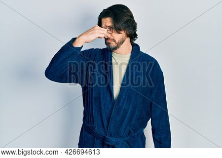 Middle age caucasian man wearing bathrobe and glasses smelling something stinky and disgusting, intolerable smell, holding breath with fingers on nose. bad smell 