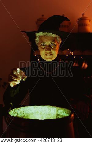 Old Witch Pouring Salt Into Her Magic Potion. Senior Person Dressed As A Witch Celebrating Halloween