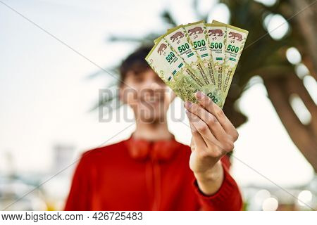 Young hispanic man using headphones holding argentinian pesos at the city