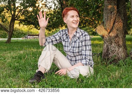 Young Woman With Short Hair Sits On Lawn In Park And Waving Her Hand In Greeting. Pleasant Meeting,