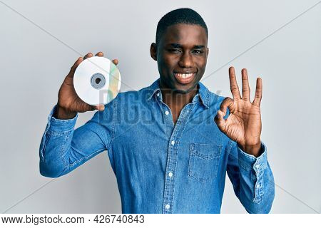 Young african american man holding compact disc doing ok sign with fingers, smiling friendly gesturing excellent symbol 