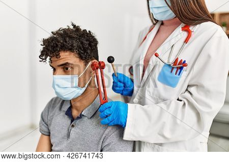 Young latin doctor woman wearing medical mask auscultating the ear of man using diapason at examination room.