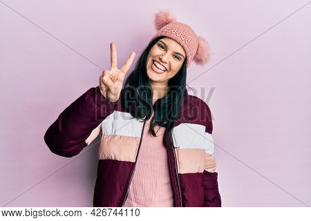 Young hispanic woman wearing wool winter sweater and cap showing and pointing up with fingers number two while smiling confident and happy. 