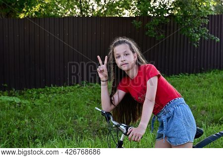 Beautiful Teenage Girl In Denim Shorts And T-shirt Is Learning To Ride Bicycle Through Streets Of Su