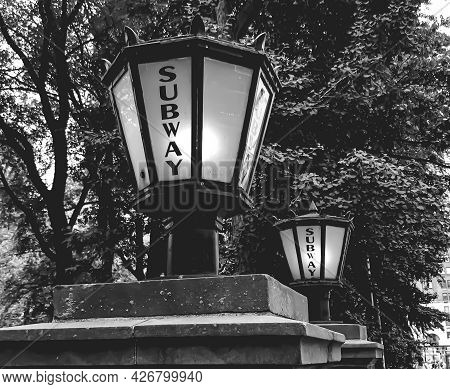 Side Angle Shot Of An Old Fashioned Lamp On A Short Pole Near Another Lamp With The Inscription 