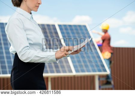 Serious businesswoman reading document when mounter installing solar panel in background