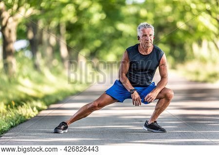 Grey-haired Old Man Still In Shape Streches Legs In His Pre-workout Warmup.