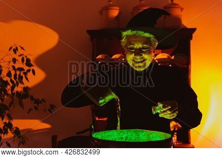 Older Woman Disguised As A Classic Witch With A Malefic Smile Preparing A Magic Potion That Emits Gr
