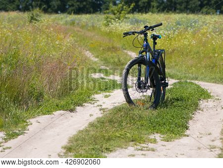 Bike Stands On The Road In The Field. A Mountain Bike Stands On A Field Path With Green Grass. Mount