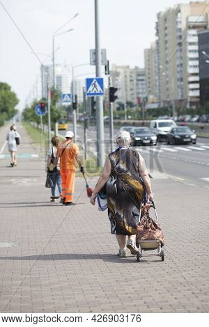 A Plump, Fat Poor Elderly Woman With A Heavy Shopping Bag Walks Through The City. Back View. 07.15.2