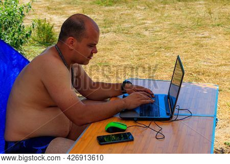 Man Using His Laptop Sitting At A Folding Picnic Table In A Camping. Working While Traveling. Freela
