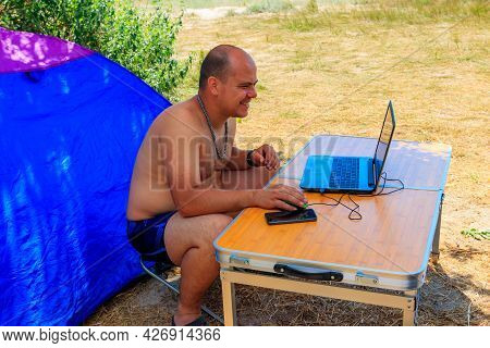 Man Using His Laptop Sitting At A Folding Picnic Table In A Camping. Working While Traveling. Freela