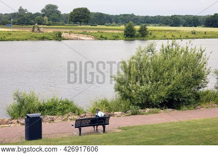 Arcen, The Netherlands - June 24, 2021: Woman Reading A Book At Bench Near River Meuse In Dutch Arce