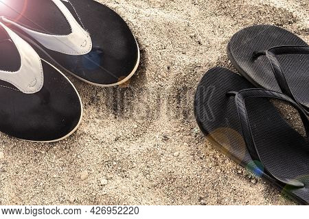 Mens And Womens Beach Shoes On The Sand Under The Rays Of The Sun. Two Pairs Of Black Flip-flops