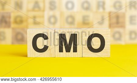The Text Cmo Is Written On Wooden Cubes On A Bright Yellow Surface. In The Background Are Rows Of Cu