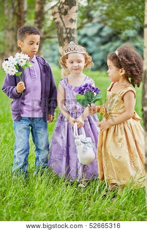 Boy and two little girls dressed in party frocks stand in park on grassy lawn, boy and one of girl hold flower