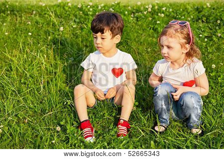 Two little children sit on grassy slope, red hearts pinned to theirs shirts