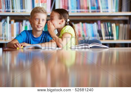 Portrait of cute schoolgirl whispering something to her classmate in library