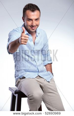 young casual man sitting on a chair and showing the thumb up gesture with a smile on his face and a hand in his pocket. on gray background