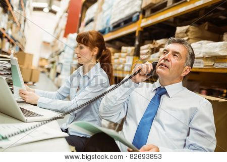 Manager working on laptop and talking on phone at desk in a large warehouse