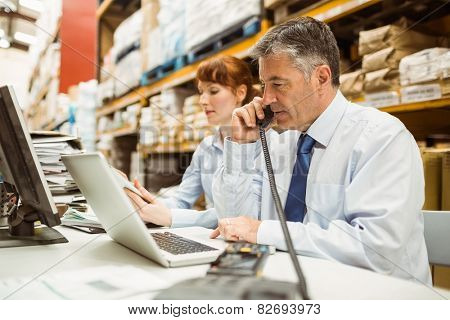 Manager working on laptop and talking on phone at desk in a large warehouse