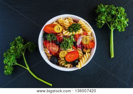 Salad: pasta fusilli black and green olives cherry tomatoes red onion and parsley. Dressing: olive oil and lemon juice. In white bowl. Black stone background. Top view.